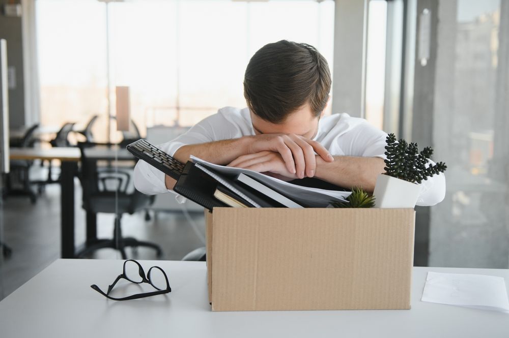 Man with head down at desk; a cardboard box with office belongings.
