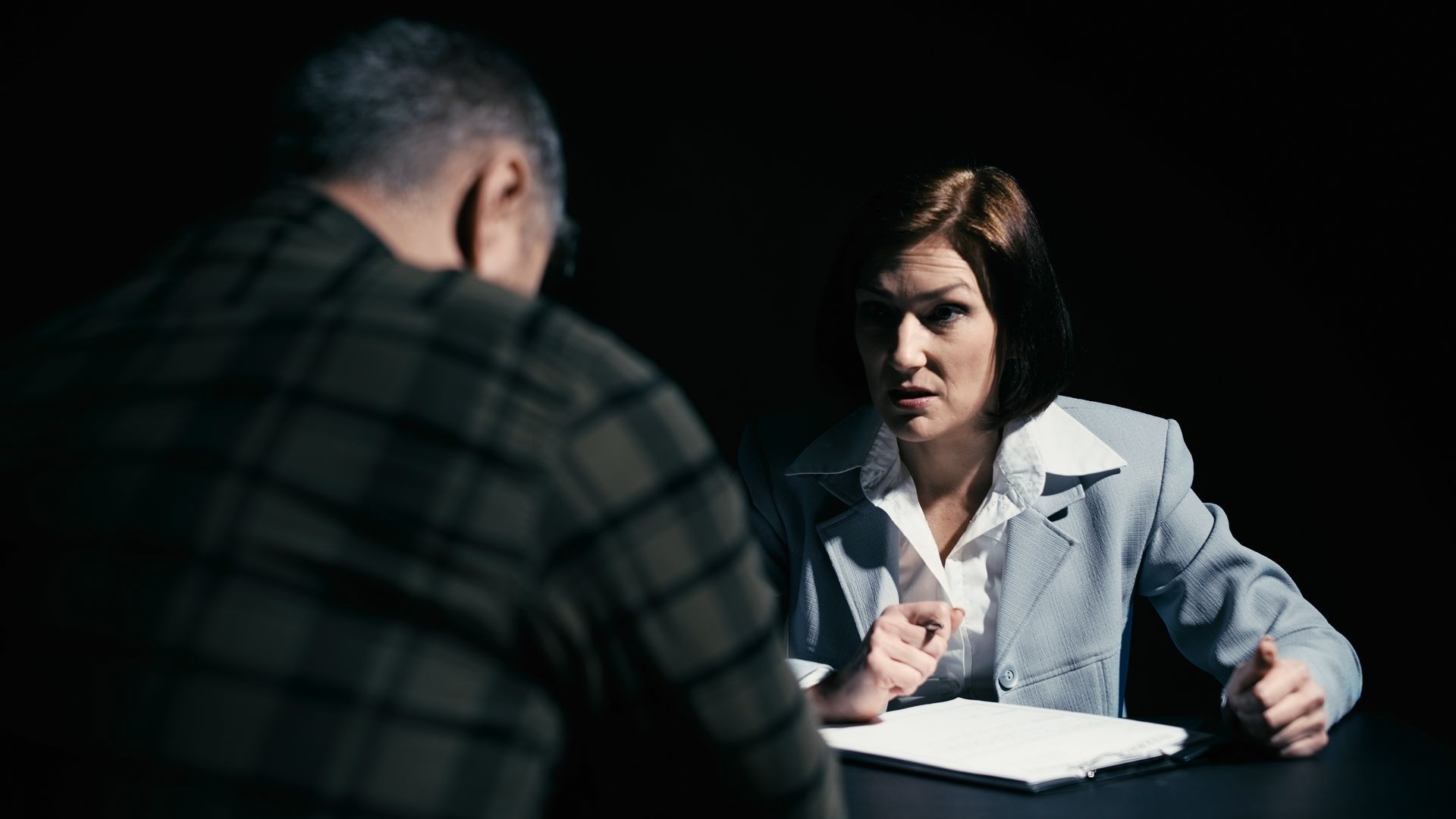 Woman in a gray suit confronts a person in a plaid shirt at a table in a dimly lit room.