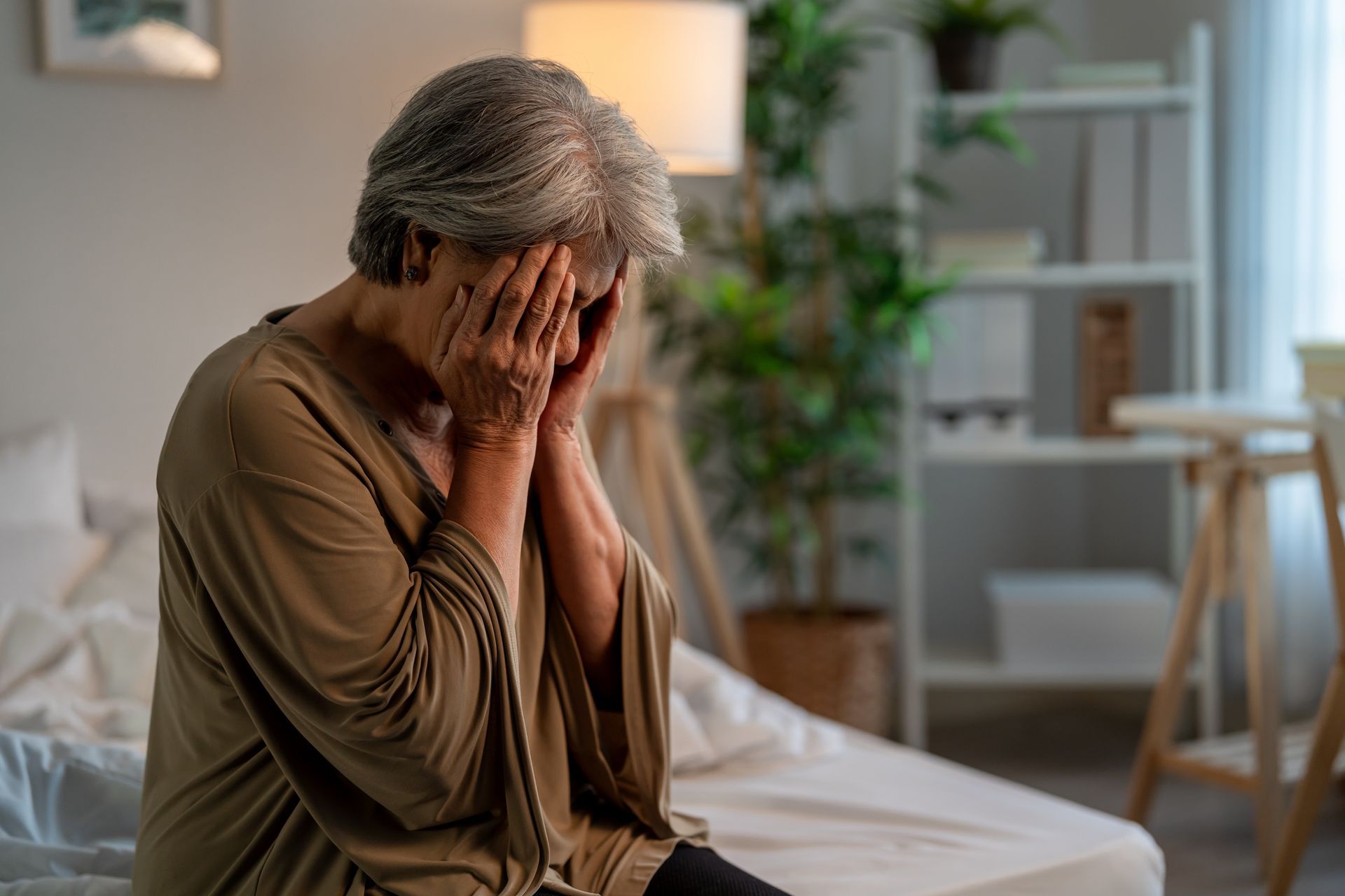 Woman sitting on a bed, hands covering her face, looking distressed.