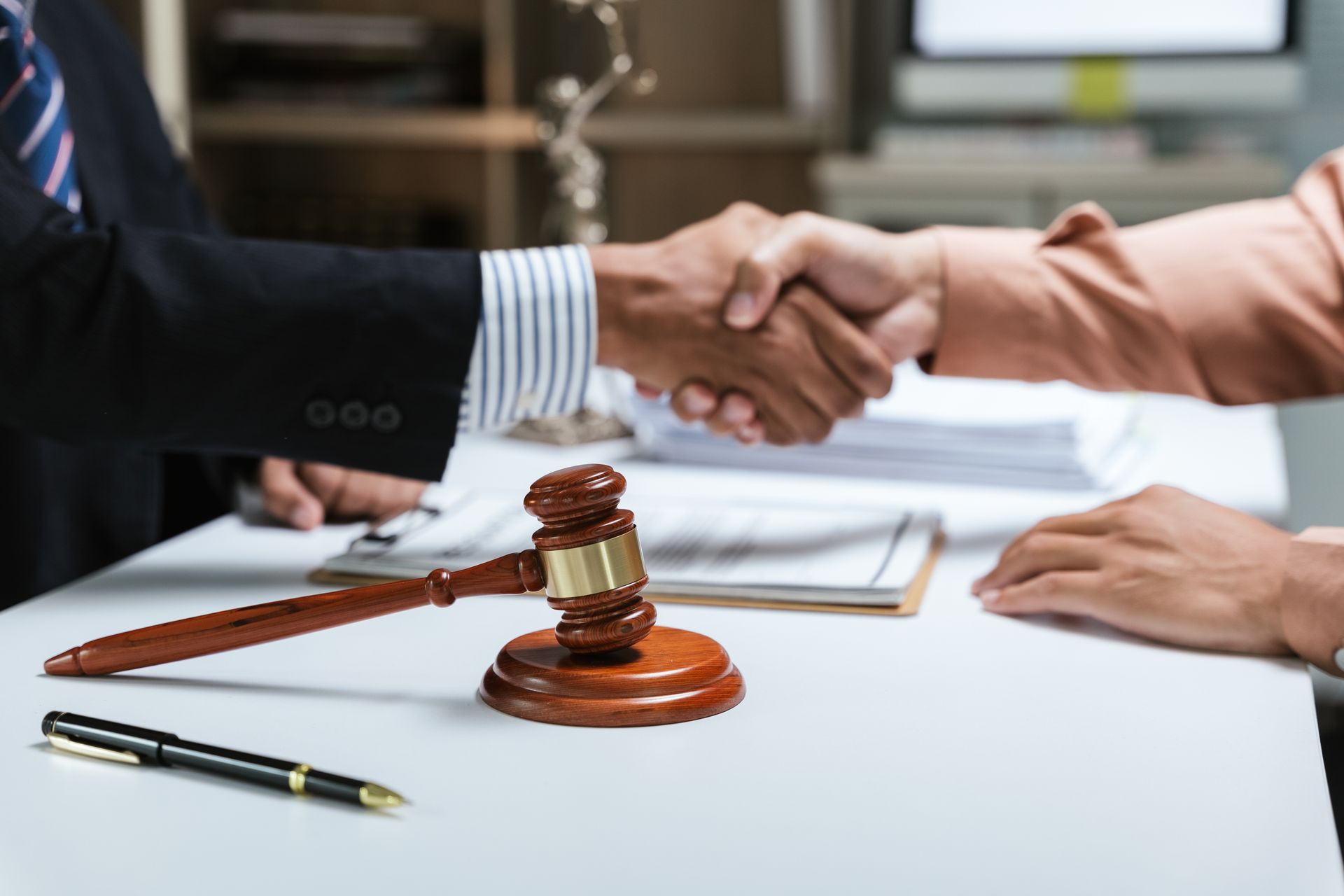 Two people shaking hands over a table with a gavel, pen, and documents.