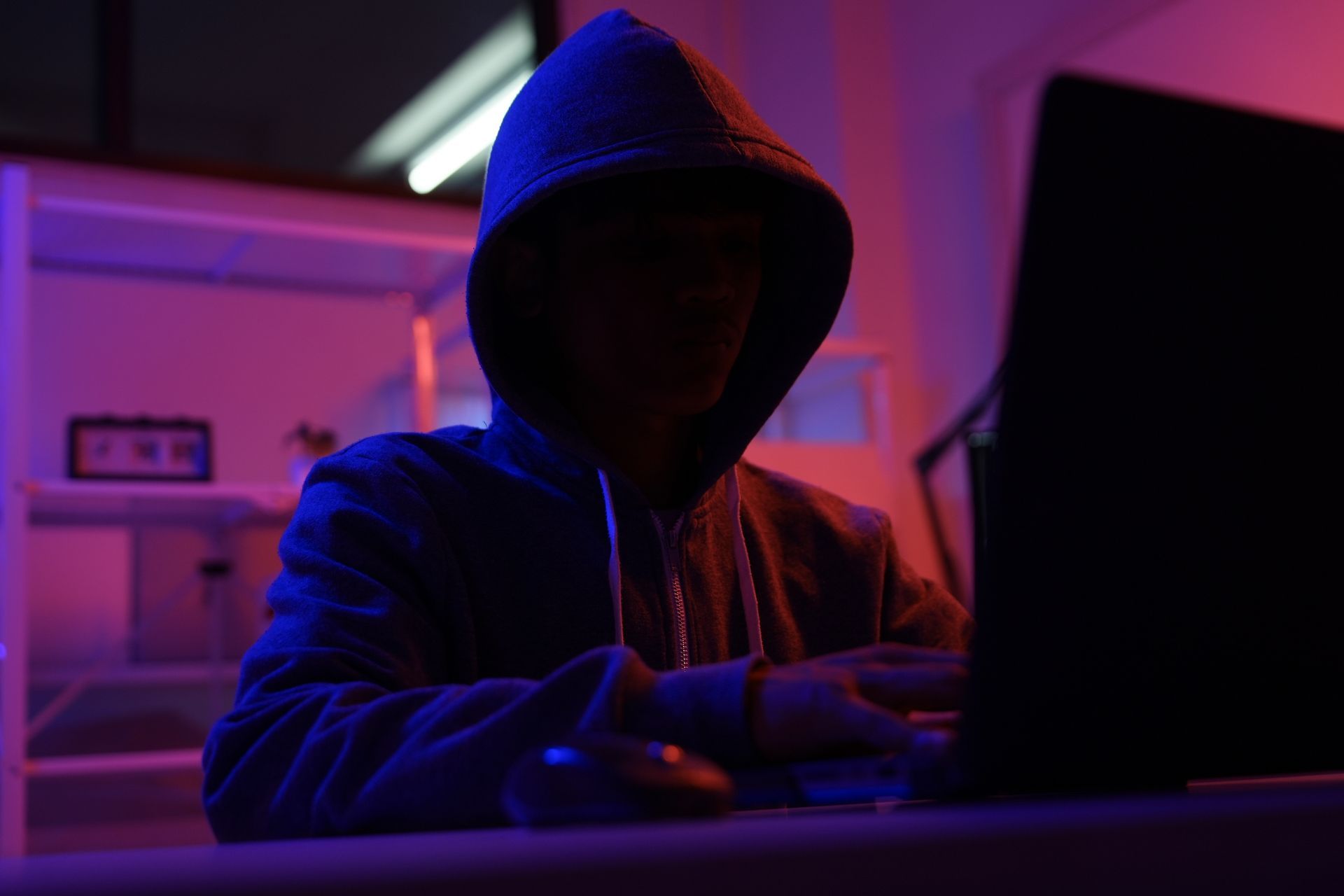 Person in hooded sweatshirt using laptop in a room lit with purple and blue light.