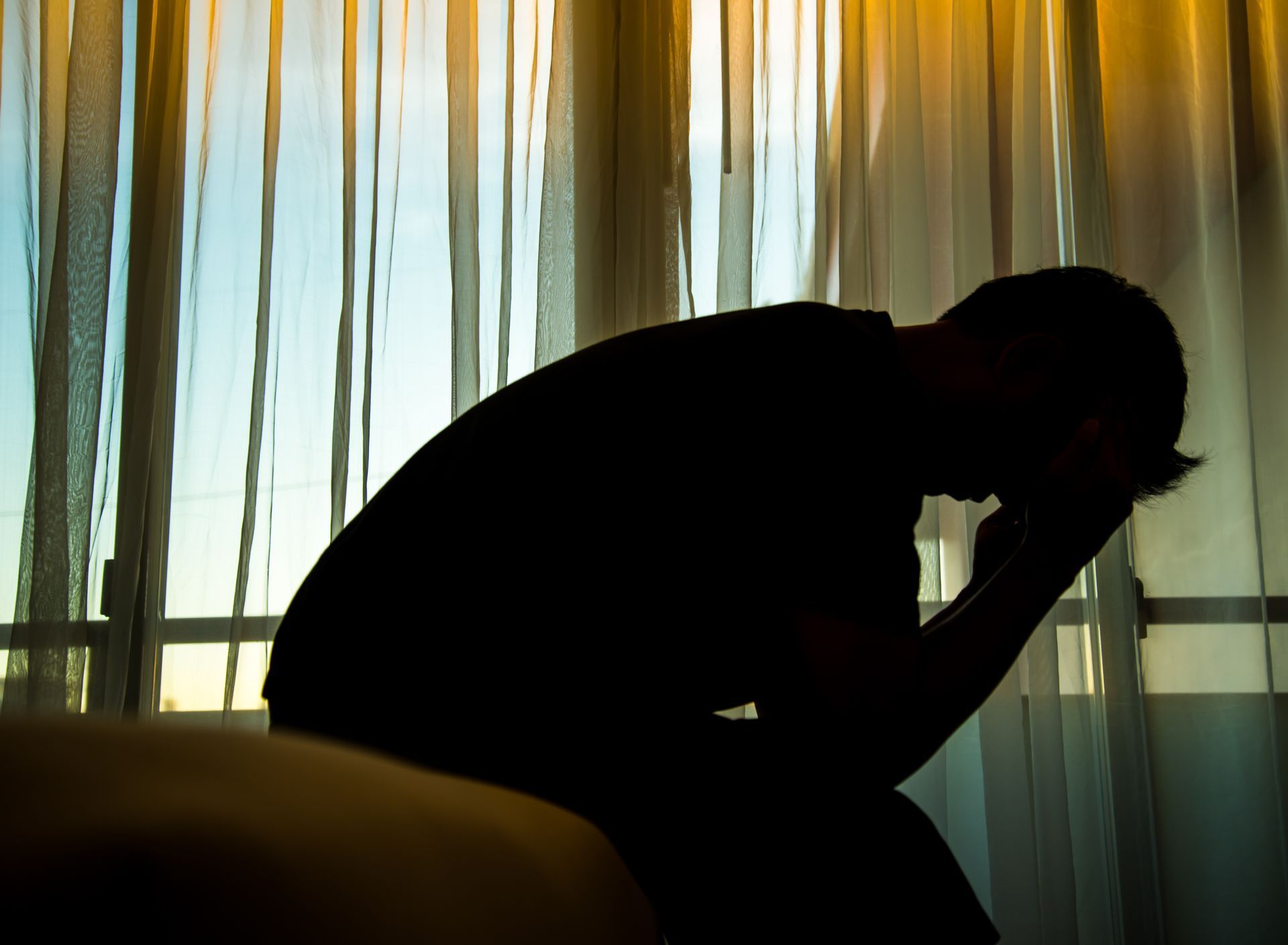 Silhouette of a person with head in hands, sitting near a window with sheer curtains.