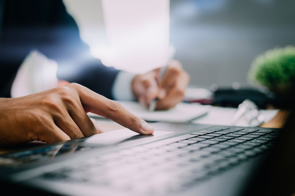 Person's hand using laptop, other hand writing in notebook, indoors.
