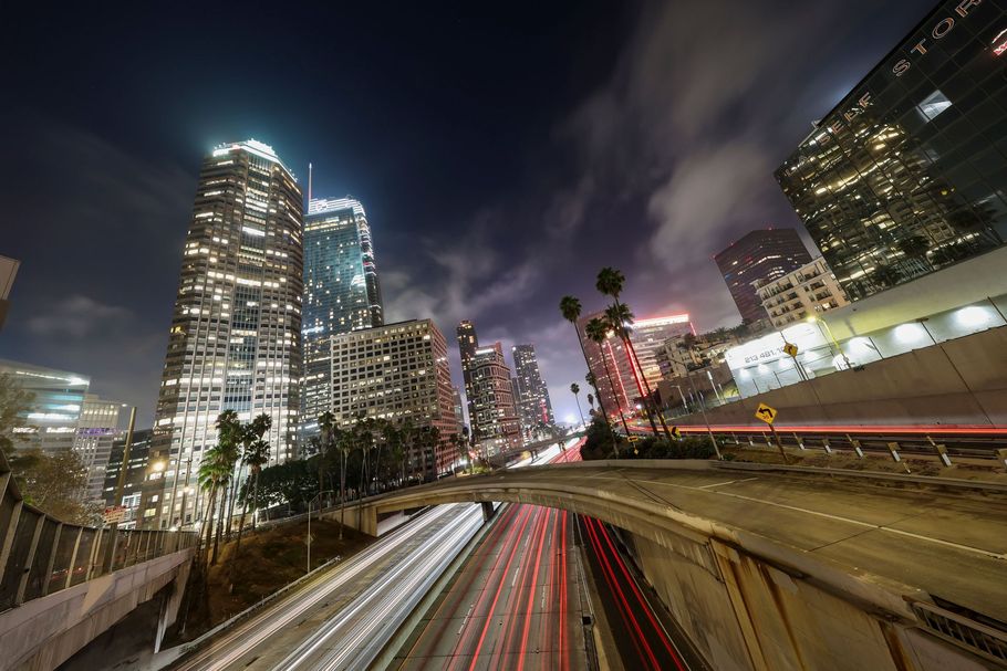 Nighttime city skyline with illuminated skyscrapers and red-white light trails on a curving highway.