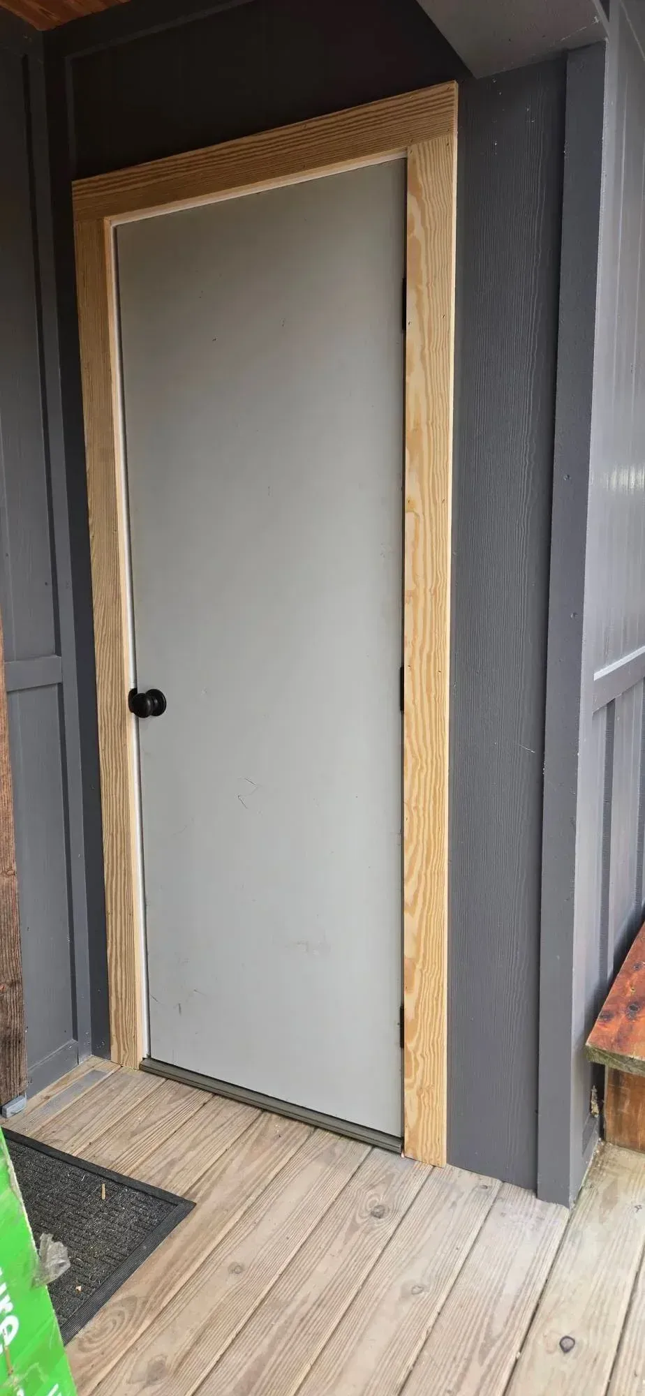 Gray door with a black knob set in a dark exterior wall, next to a wooden deck.