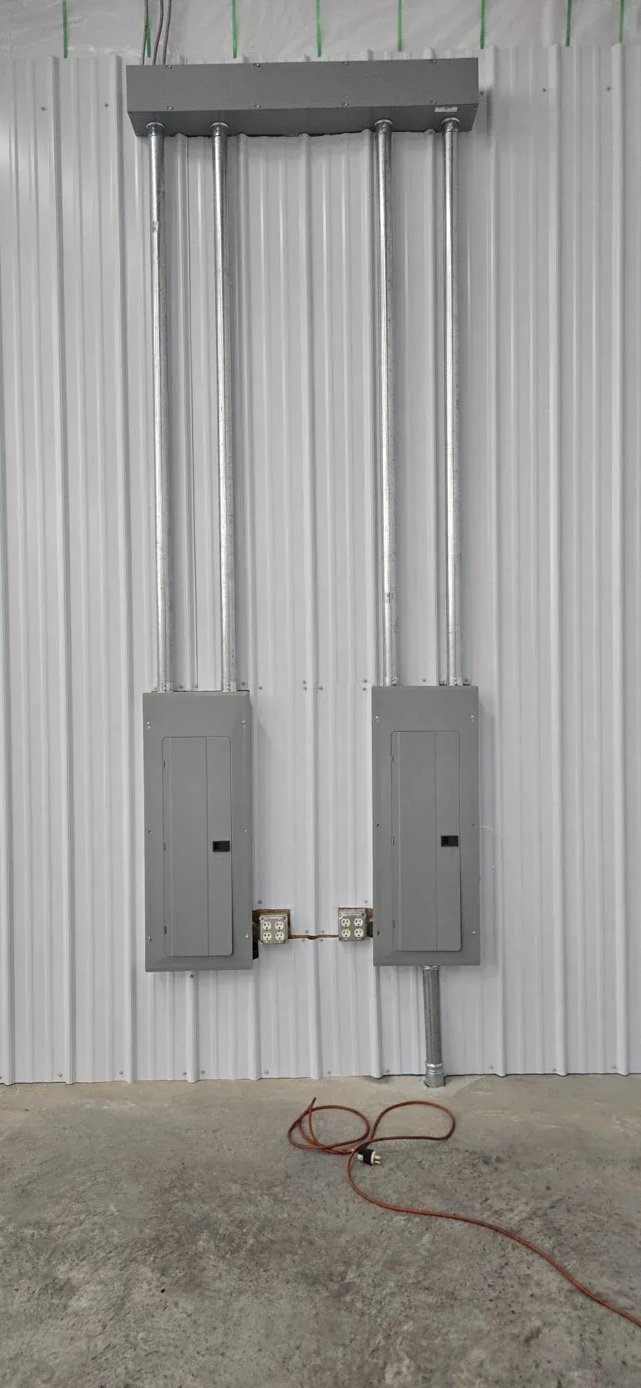 Two gray electrical boxes mounted on a corrugated metal wall with exposed conduits and a coiled cable below
