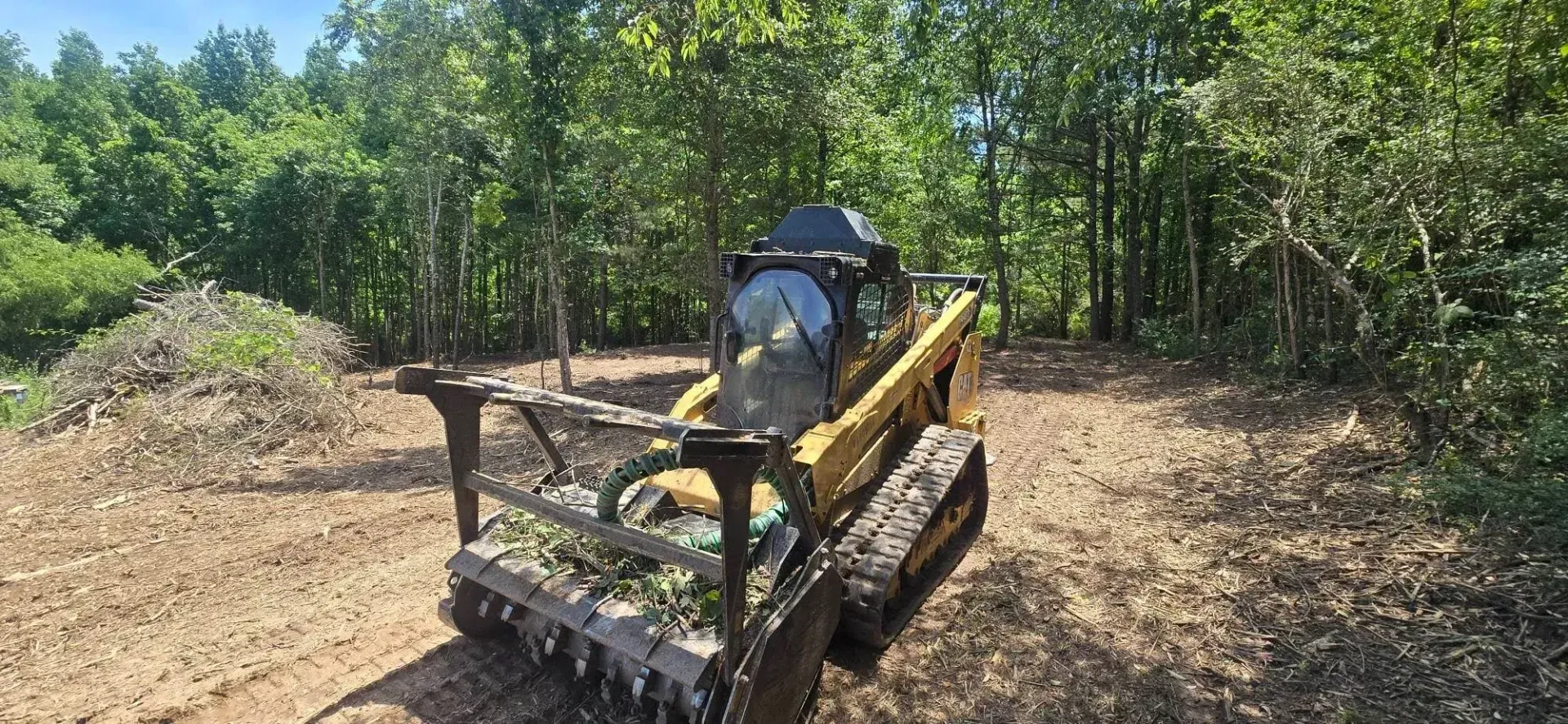 Yellow skid steer in a cleared wooded area, pushing dirt with a front attachment.