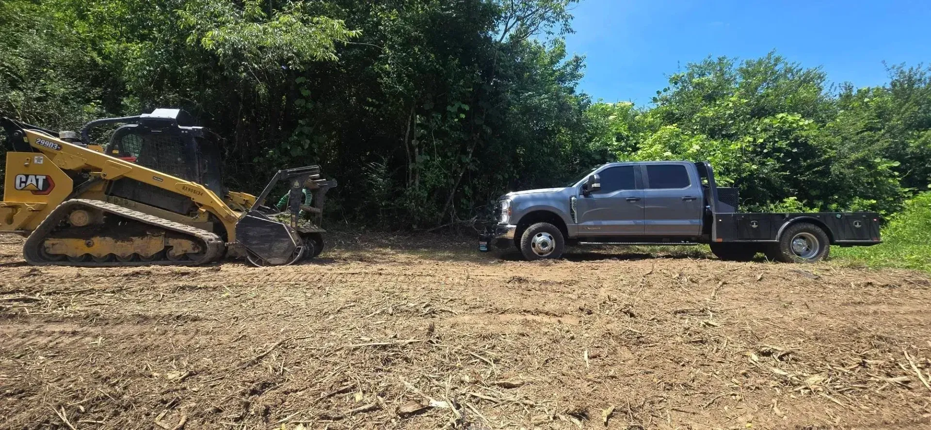 Yellow excavator and black pickup truck on a cleared dirt lot beside trees under a bright sky