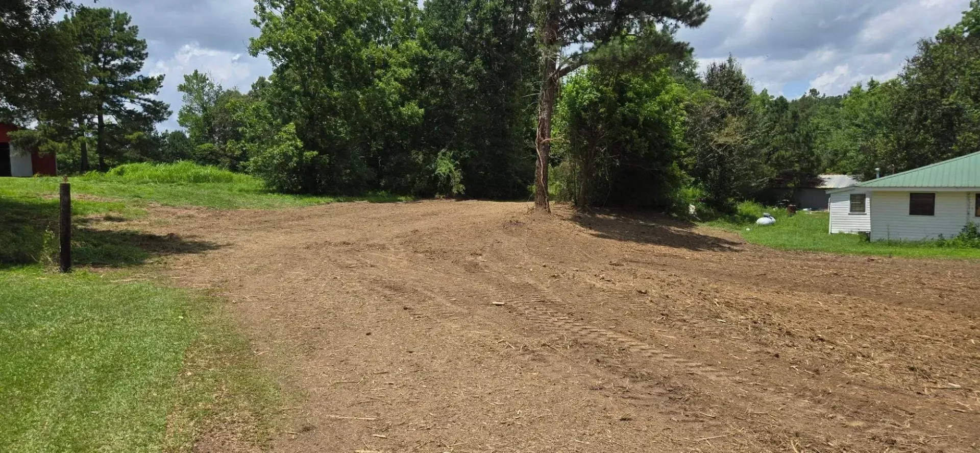 Freshly graded dirt lot beside a grassy roadside, with trees and a small house under cloudy skies
