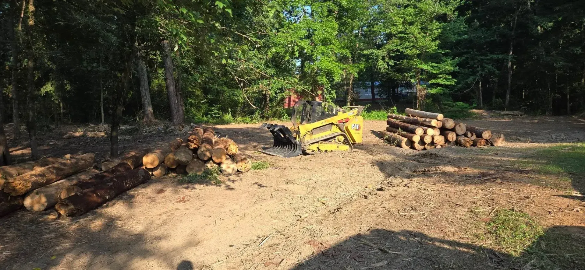 Pile of cut logs and firewood near a yellow tractor in a wooded yard