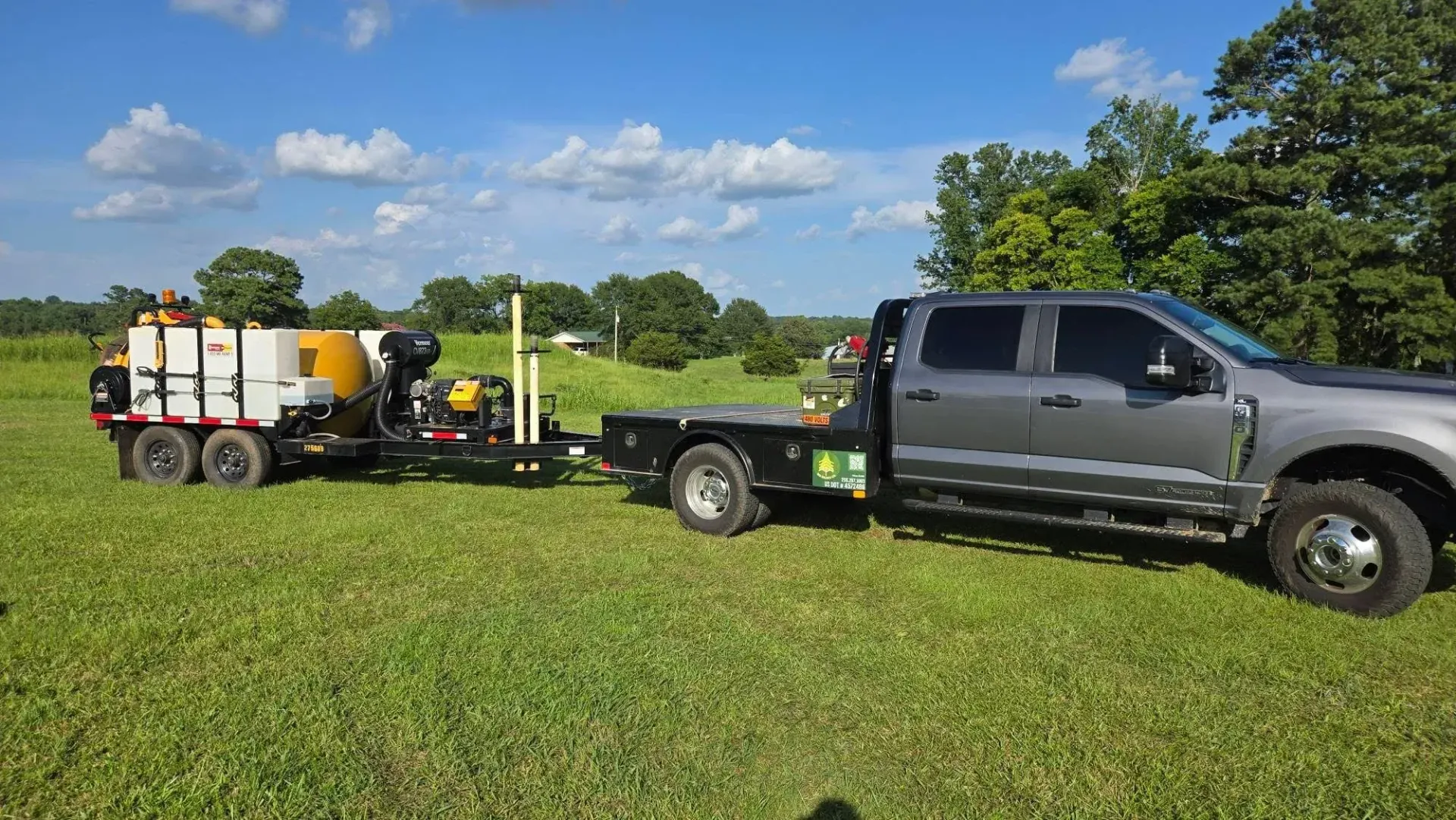 Gray pickup truck towing a utility trailer with tanks and equipment across a green field under a blue sky