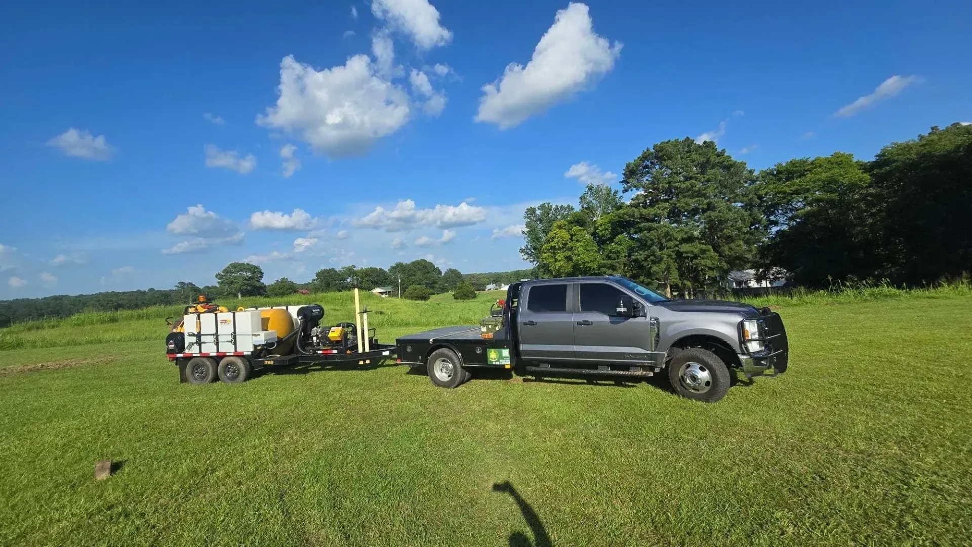 Gray pickup truck towing a small trailer with tanks across a grassy field under a blue sky