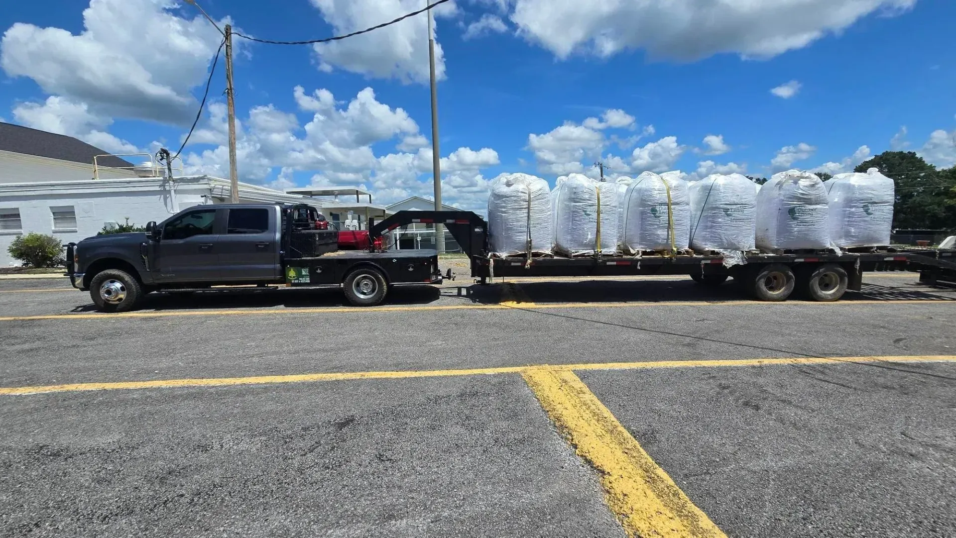 Black pickup hauling a flatbed trailer loaded with white wrapped cargo under a blue sky.
