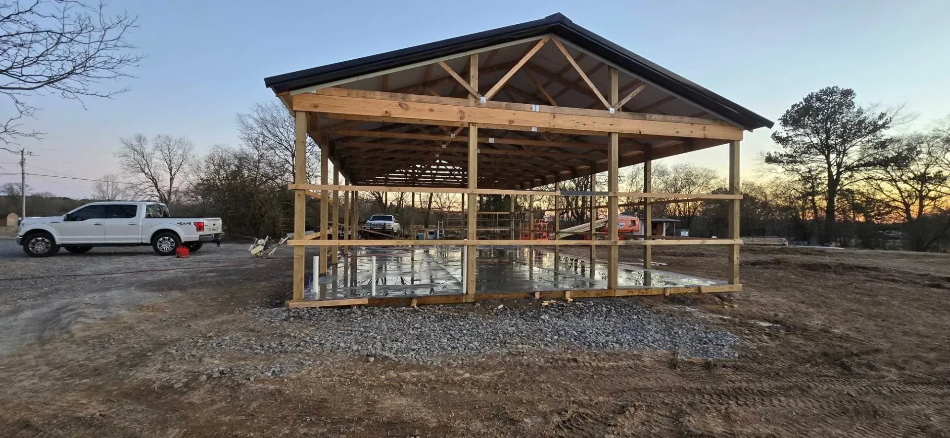Open-sided wooden pavilion on a gravel lot at sunset, with a white pickup truck nearby.