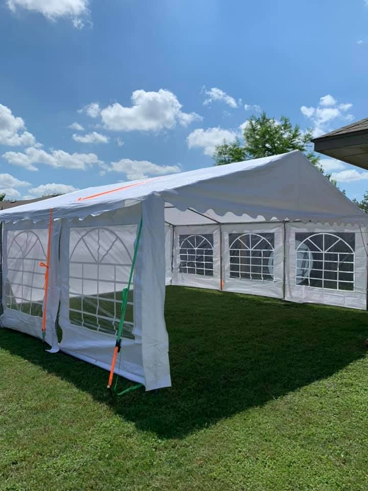 A white event tent with arched windows and orange tie-down straps stands on a green grass lawn under a blue sky.