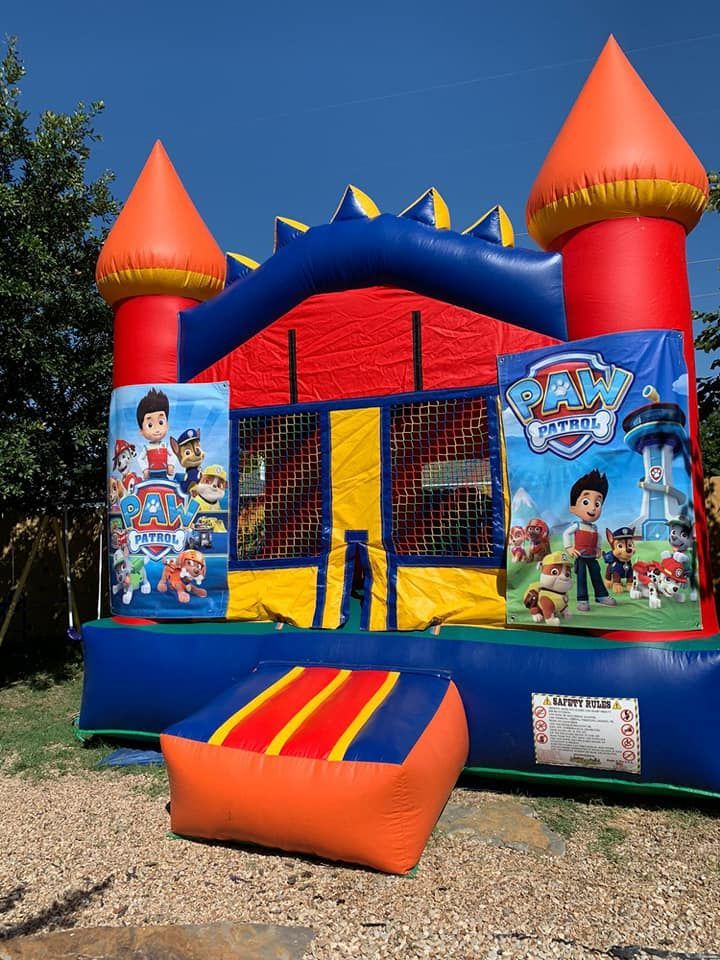 A Paw Patrol-themed inflatable bounce house with red, blue, and orange colors set outdoors on a gravel ground.