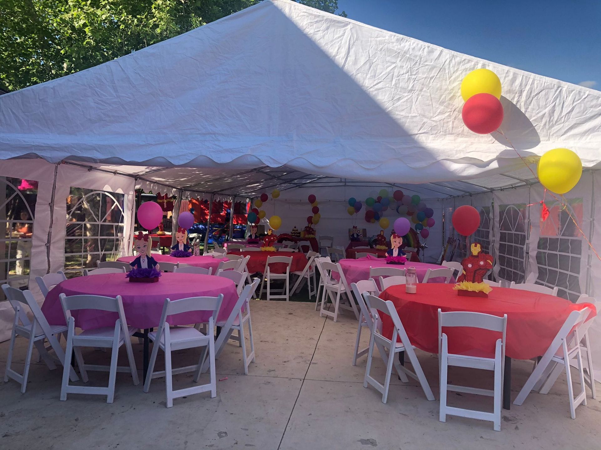 A white party tent featuring round tables with bright pink and red tablecloths, white folding chairs, and balloons.