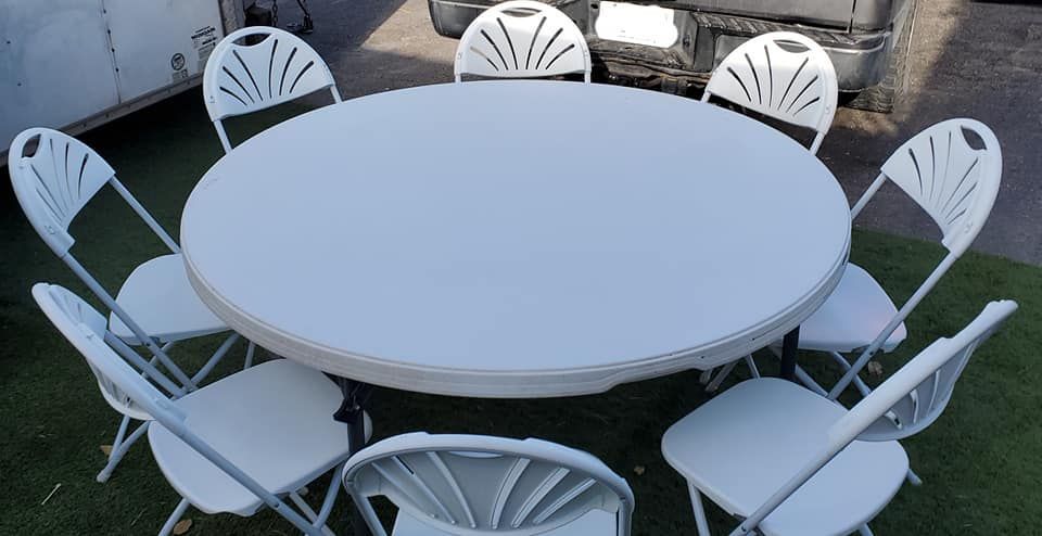 A round, white folding table surrounded by eight matching white folding chairs on a lawn outdoors.