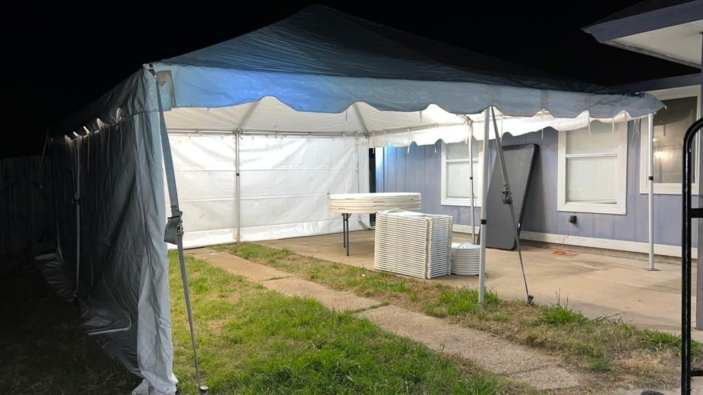 A large, white event tent set up on a grassy lawn next to a light blue house at night, illuminated by interior lights.