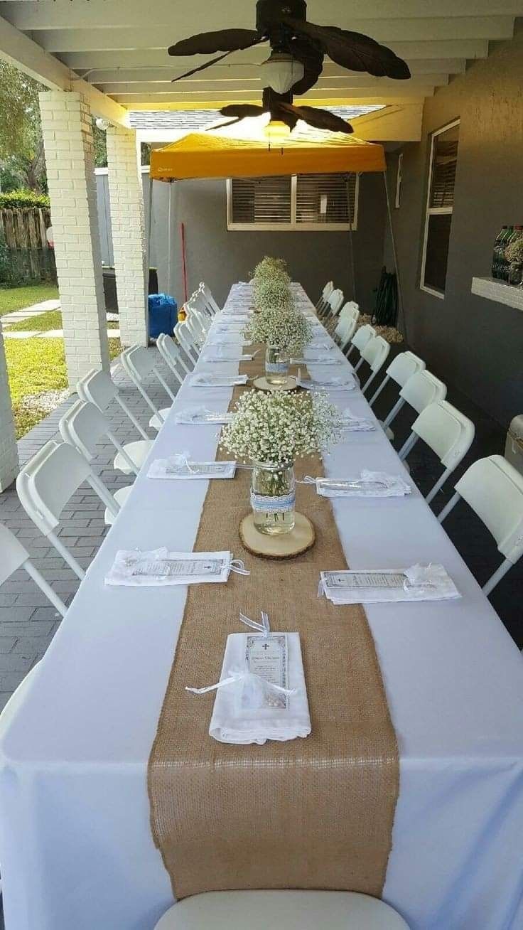 A long banquet table set for a rustic event, featuring a burlap runner, baby's breath centerpieces, and white chairs.