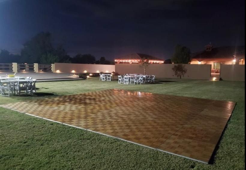 An outdoor nighttime setting featuring a large, wooden parquet dance floor on a grass lawn with dining tables in the back.