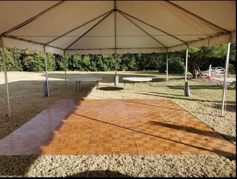 A large square wooden dance floor sits under a white event tent on a grass lawn with two tables in the background.