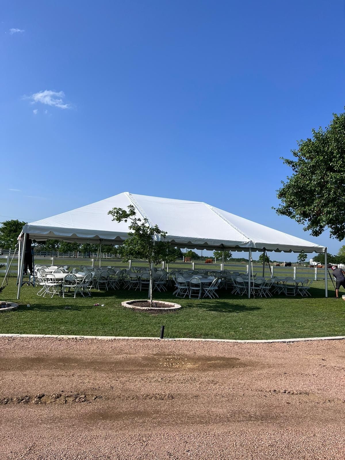 A white event tent set up on a grassy lawn with tables and chairs underneath, viewed from a gravel area under a blue sky.