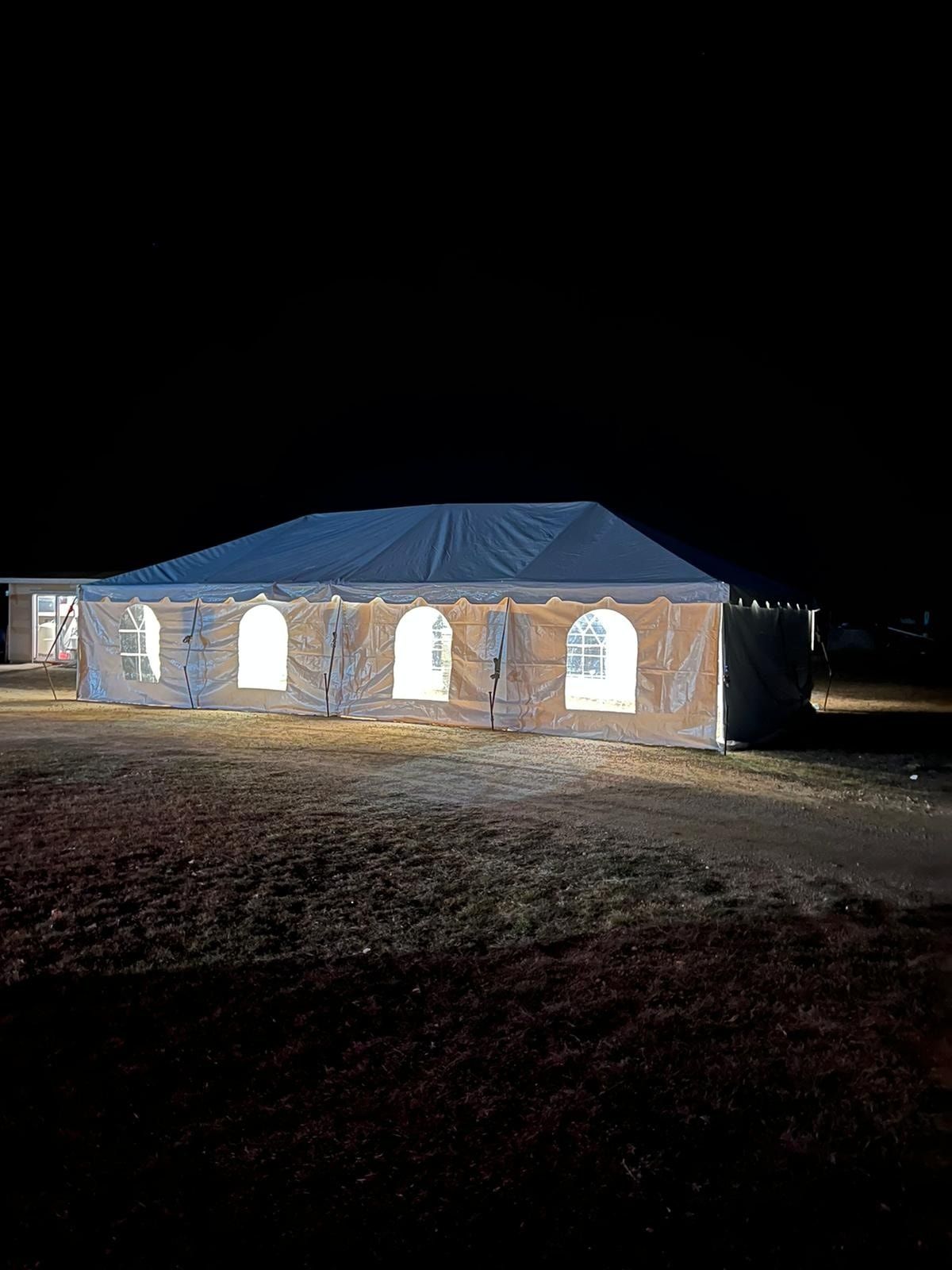 A large white event tent with illuminated arched windows stands on a dark, grassy field at night.
