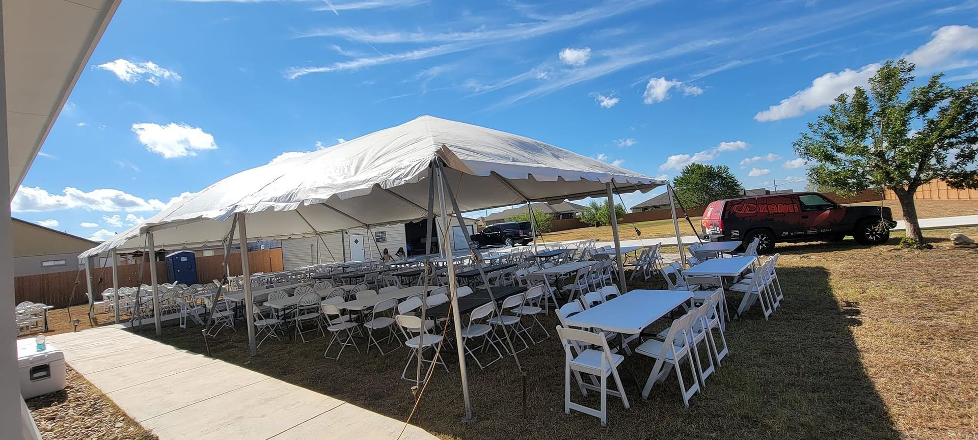 Rows of white folding tables and chairs set up under a large white party tent in an open, gravel-covered outdoor area.