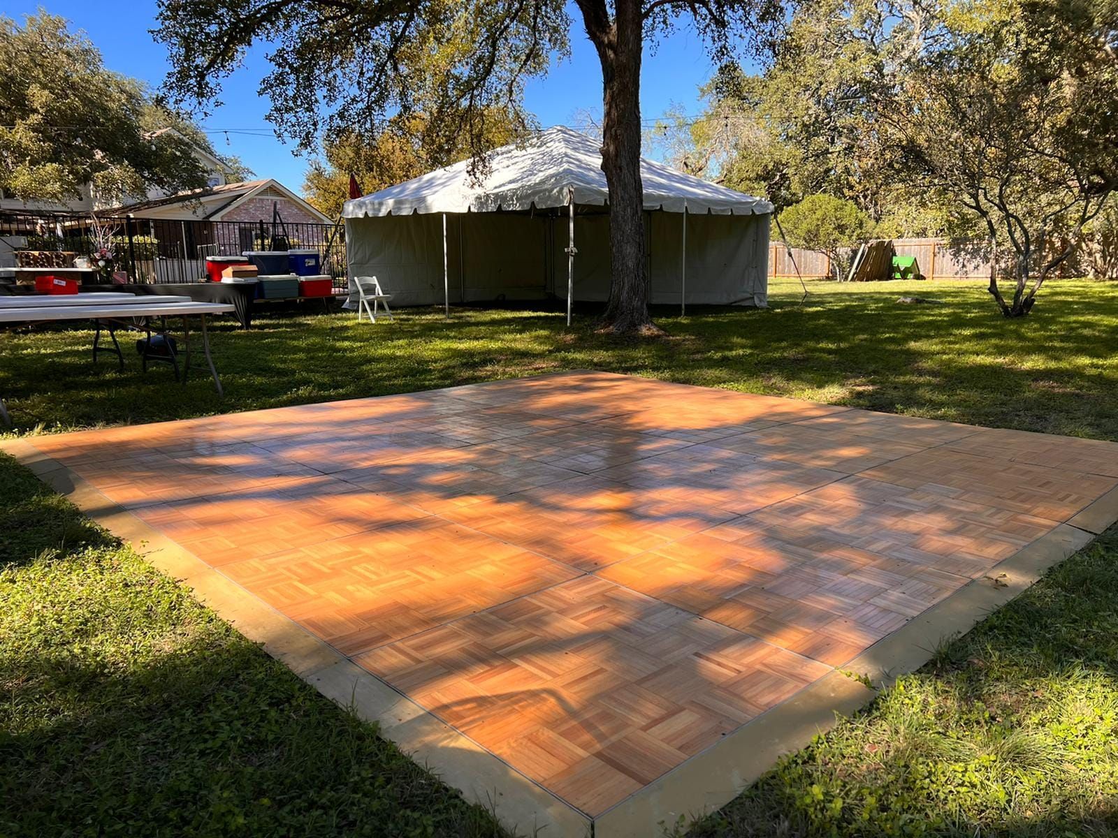 A square, portable wooden dance floor set up on a grassy lawn outdoors near a large white tent under a clear blue sky.