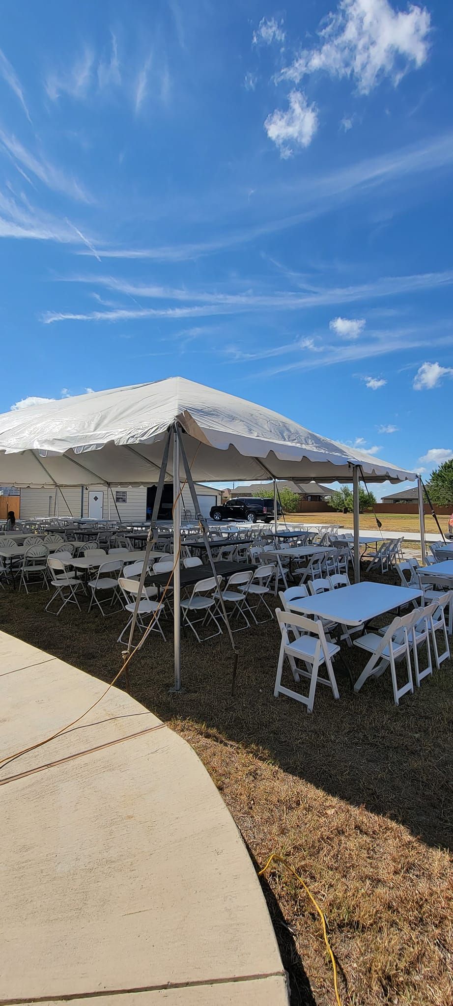A white event tent stands over rows of tables and chairs set up on a gravel lot under a bright, sunny blue sky.