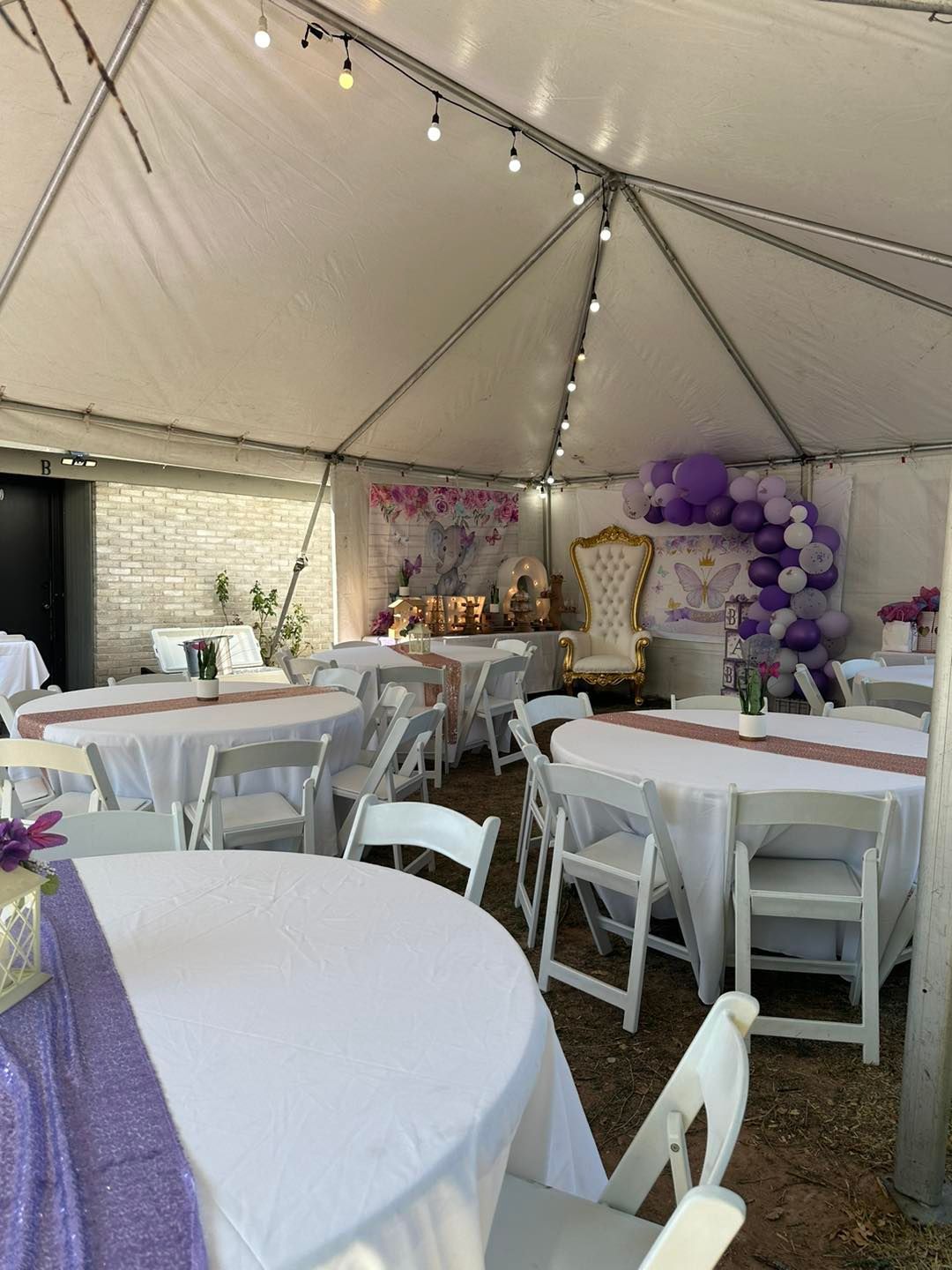 A white tent set up for a party with round tables, white chairs, purple balloon arches, and a gold decorative chair.