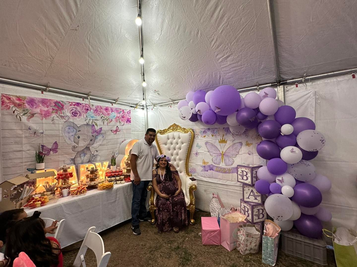 A person stands beside a seated person in a dress at a baby shower with floral decor, a throne chair, and balloon arch.