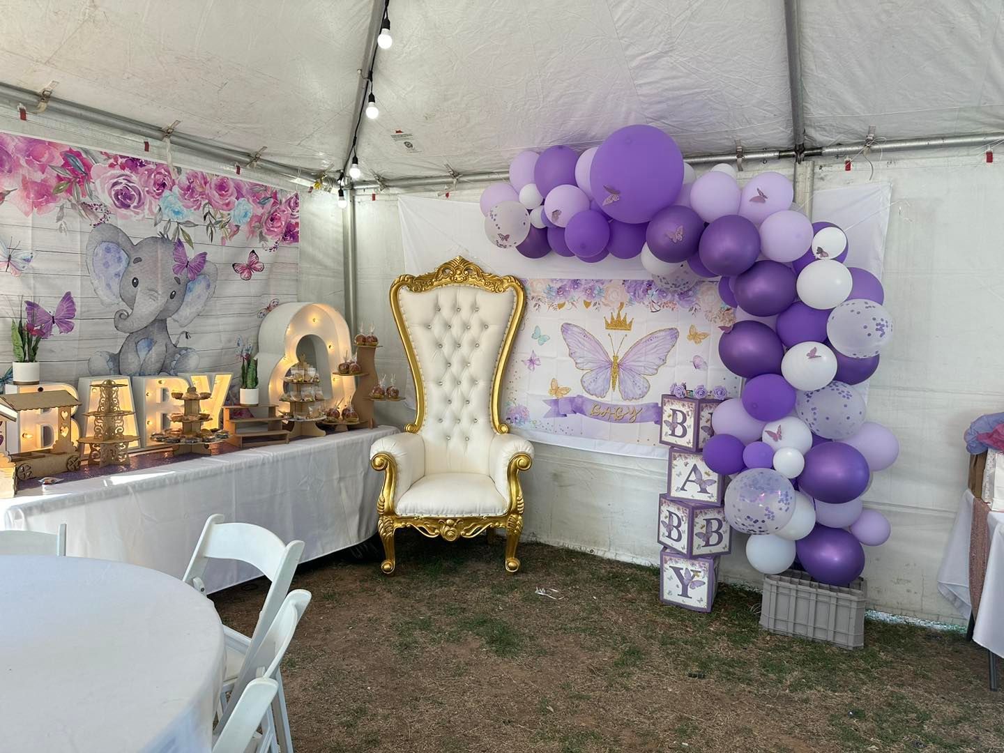 A baby shower tent with a white throne chair, purple and white balloon arch, 
