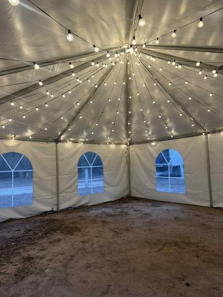 A white event tent interior lit by strings of hanging lights, featuring arched windows and a dirt floor.