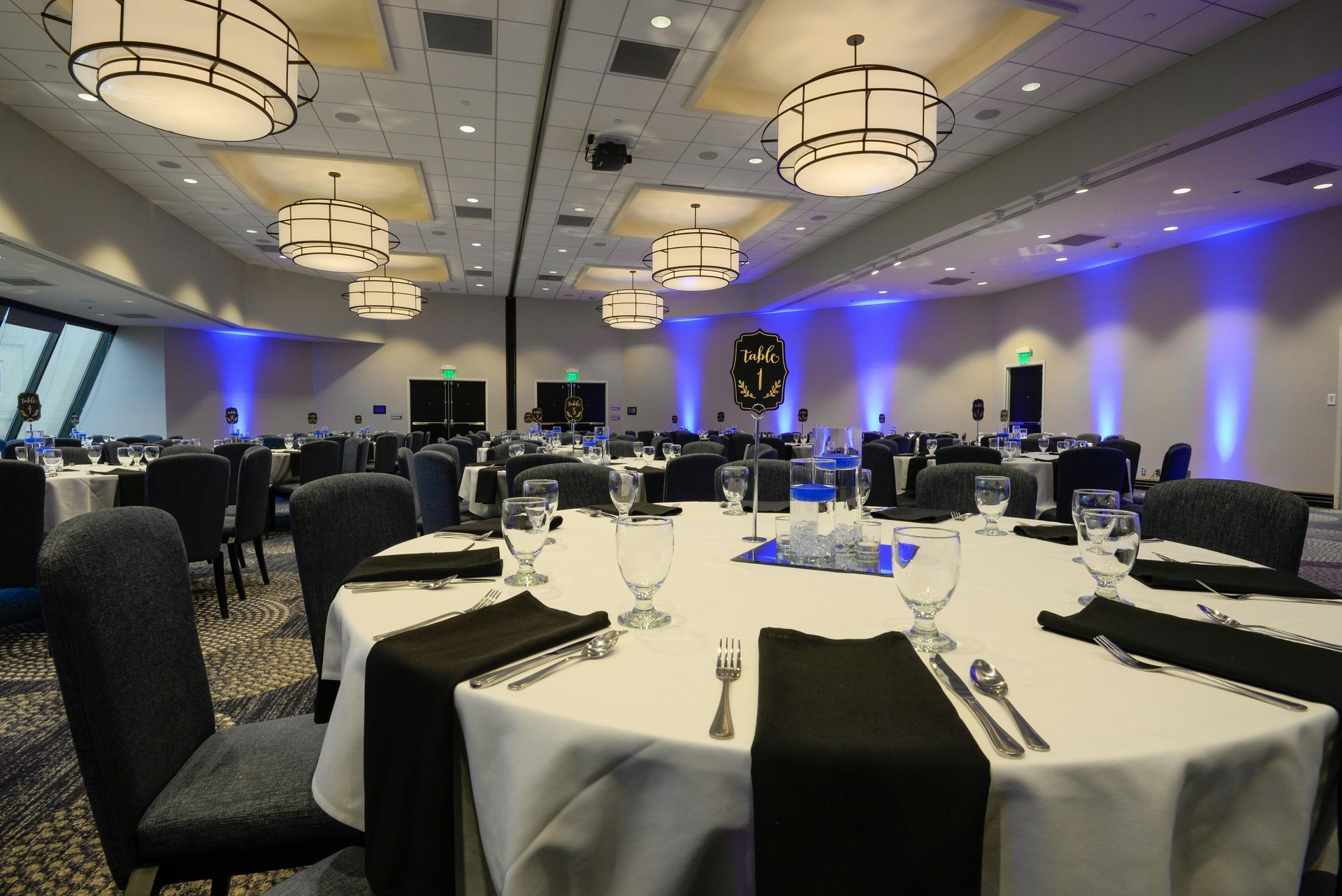 A banquet hall set for a formal event with round tables covered in white tablecloths and dark chairs under pendant lights.