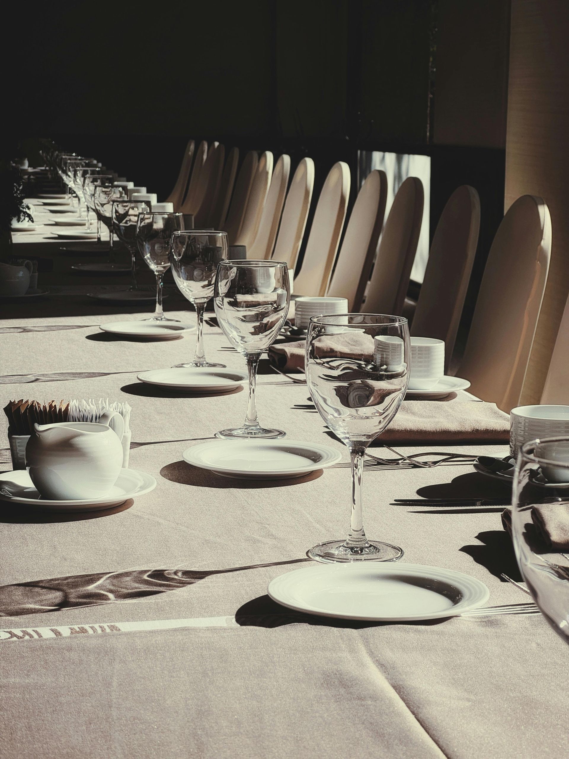 A long, formal dining table set for many guests, with wine glasses, plates, and chairs lined up in soft, natural light.