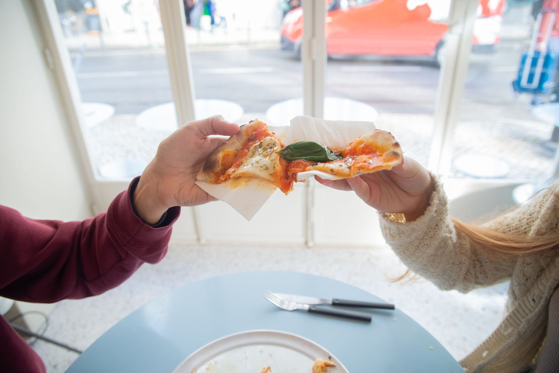 Two people clink pizza slices over a table in a bright cafe by a window.