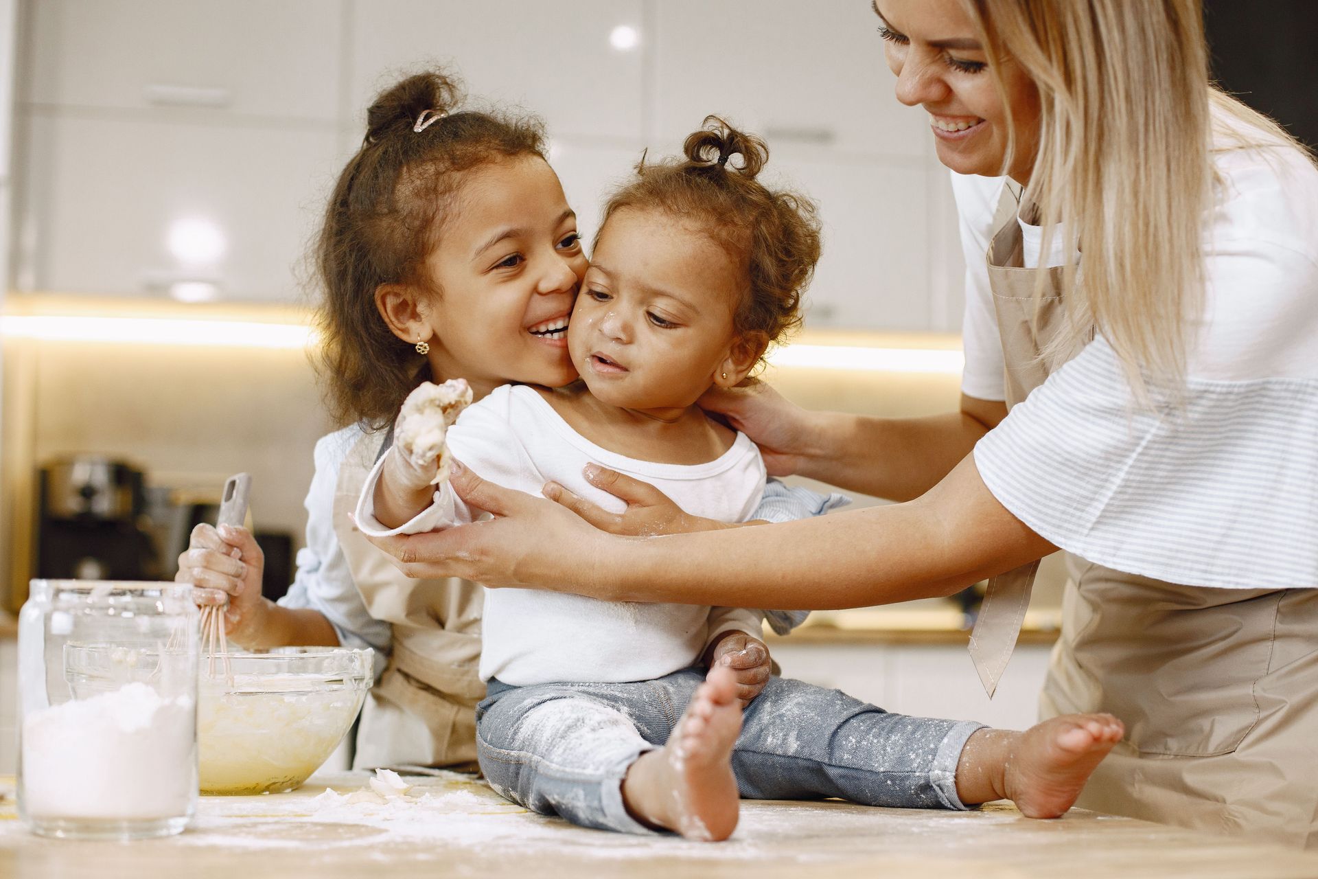Adult and two children baking together in a bright kitchen, smiling by a countertop with ingredients.