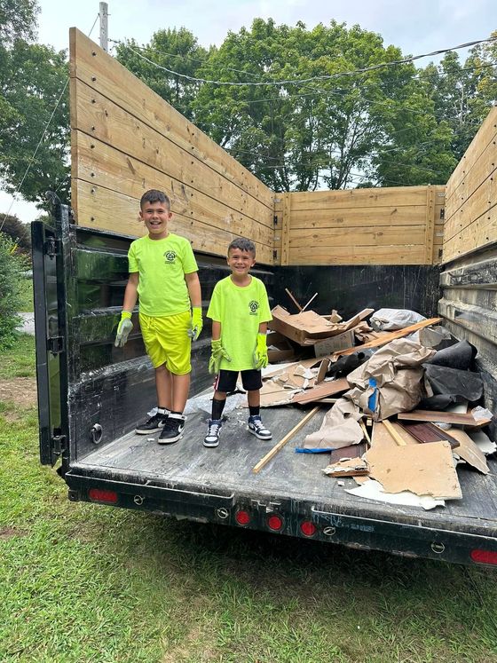 Two young boys are standing in the back of a dumpster filled with cardboard.