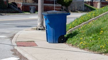 A blue trash can is sitting on the sidewalk next to the street