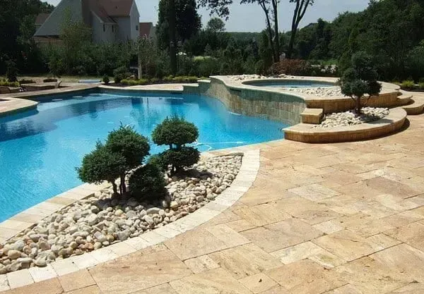 Pool and spa with tan stone patio, manicured shrubs, and clear blue water. Sunny outdoor setting.