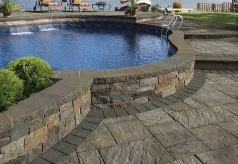 Stone-edged swimming pool with patio, steps. Landscaping with bushes and view of a lake in background.