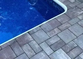 Pool edge with blue water, surrounded by gray and tan paving stones in a rectangular pattern.