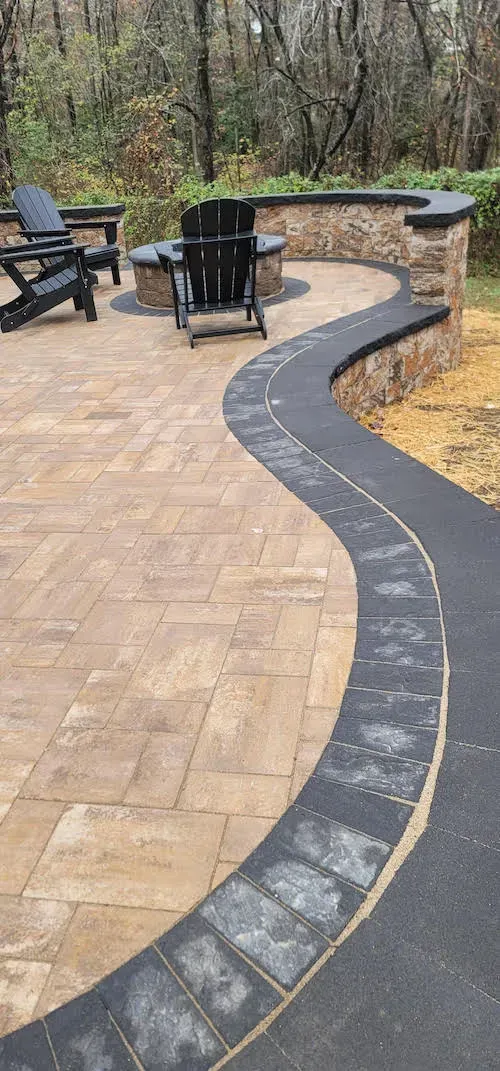 A curved patio with a fire pit, black chairs, and a dark stone border. Brown and tan pavers.