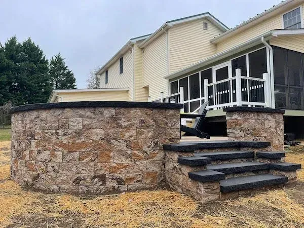 Stone-walled outdoor hot tub with steps, next to a house with a screened porch on a cloudy day.