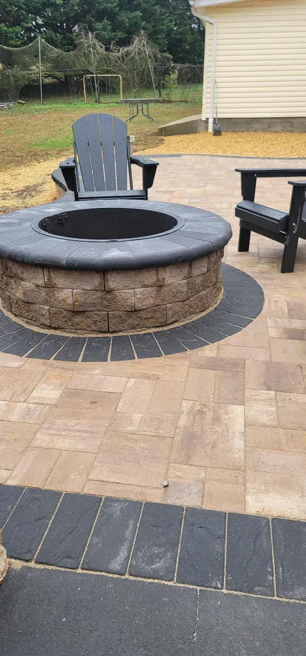A stone patio with a fire pit, surrounded by black chairs. Trees and a building are in the background.