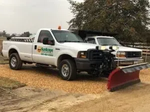 Two white trucks, one with a snowplow, parked on gravel.