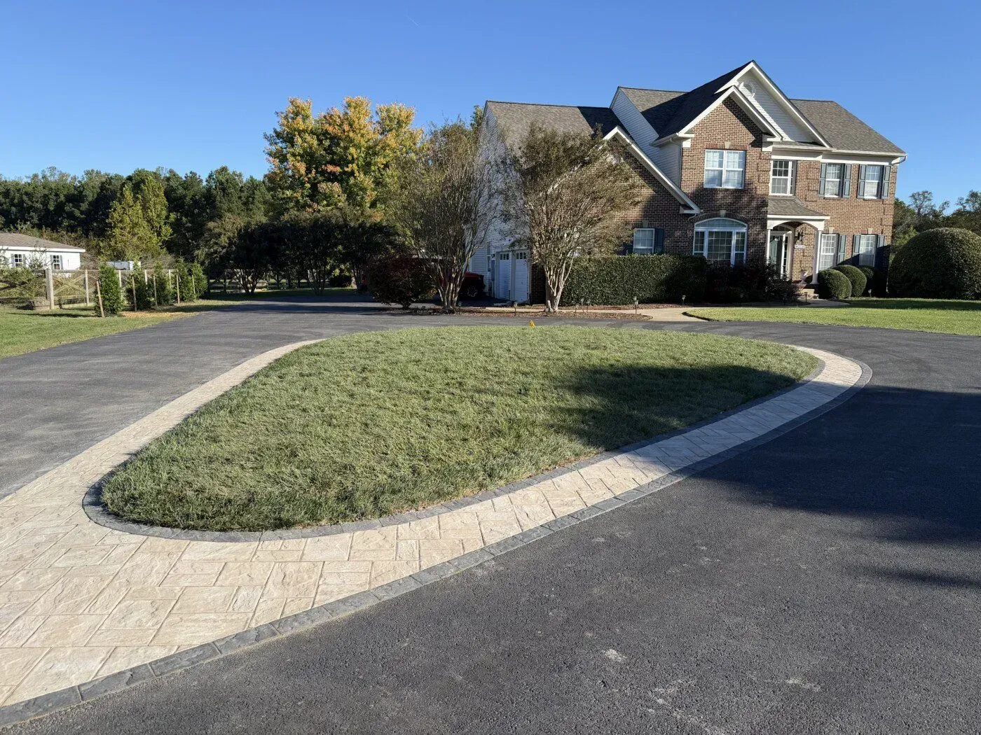 House with circular driveway, featuring a grassy island bordered by beige pavers. Blue sky, fall trees.