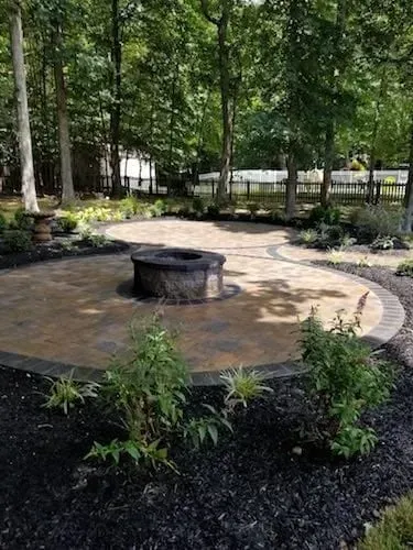 Fire pit on a circular patio surrounded by trees and plants. Dark mulch and brick border.